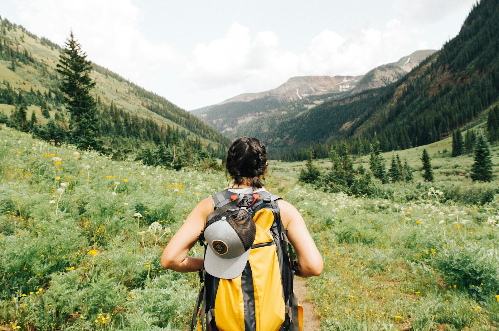 person carrying yellow and black backpack walking on a hillside. mountains are in the background.
